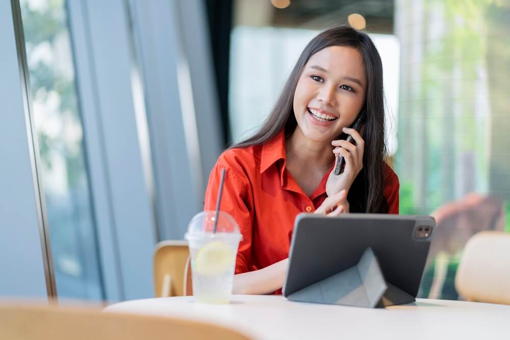 an female Asian is talking on a phone and using her tablet relaxing and happily as she is applying for hiring teachers on a website for ignite a star.